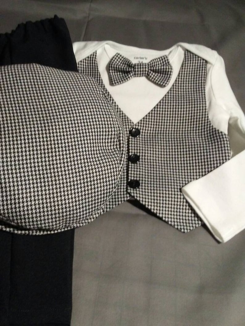 May include: A black and white houndstooth pattern baby outfit with a white long-sleeved shirt, a vest, black pants, and a matching hat. The shirt has a bow tie and the text "Carter's" on the front.