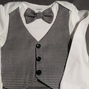 May include: A white onesie with a black and white houndstooth vest and bow tie. The vest has four black buttons. The onesie has the text "carter's" and "24" printed on the front.