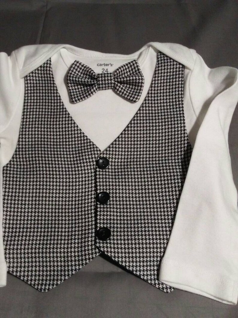 May include: A white onesie with a black and white houndstooth vest and bow tie. The vest has four black buttons. The onesie has the text "carter's" and "24" printed on the front.