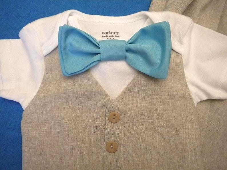 K&ouml;nnte beinhalten: Ein wei&szlig;er Strampler mit einer hellblauen Fliege und einer beigefarbenen Weste mit zwei braunen Kn&ouml;pfen. Die Weste hat einen strukturierten Stoff.