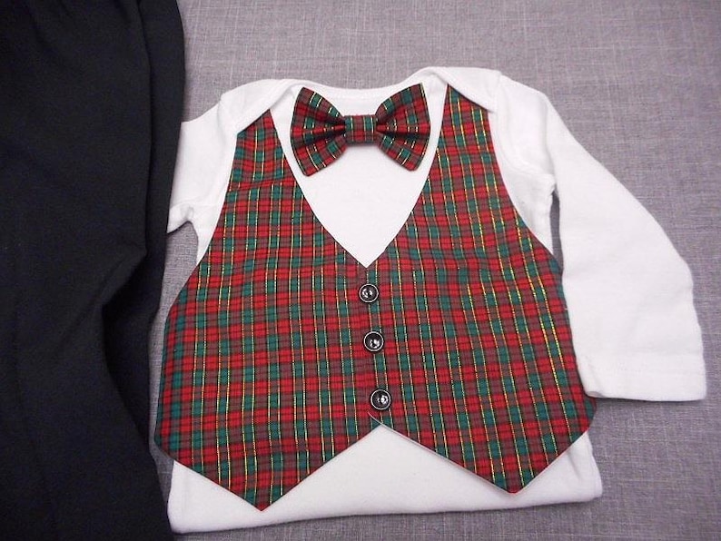 May include: A white baby bodysuit with a red, green, and gold plaid waistcoat and bow tie. The waistcoat has three black buttons. Black trousers are visible on the left. The outfit is designed for a baby.