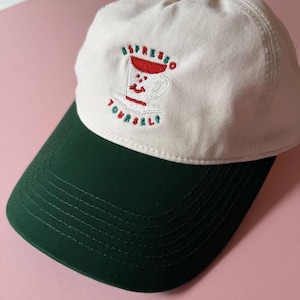 May include: A white baseball cap with a dark green brim. The front features an embroidered graphic of a smiling coffee cup with red and green details, accompanied by the text "ESPRESSO YOURSELF" stitched in red and green thread.