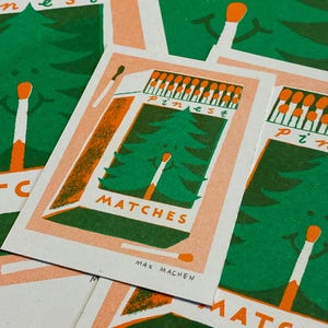 May include: A vintage-style illustration of a matchbox with a green pine tree and the text "Pines" and "Matches". The matchbox is orange and white with the text "Max Machen" at the bottom.