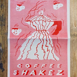 May include: A pink poster with a red and white illustration. The central image is a white figure with red lines, resembling a coffee maker. Three smiling coffee cups are scattered around the figure. The words "COFFEE SHAKEZ" are printed in red and white.