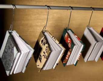 May include: Five small, bound books hang from a metal rod. The books are covered in different patterned fabrics, including floral, geometric, and solid colors. The books are all closed and appear to be handmade.