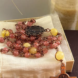 May include: A rosary with pink and yellow beads, copper wire, and a decorative metal cross. The rosary is displayed on a small box with a cream-colored cloth.