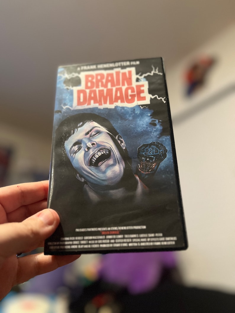 May include: A vintage VHS tape for the horror film "Brain Damage." The cover art features a screaming man with a grotesque creature emerging from his head, set against a dark, stormy background. The title is in bold red letters.