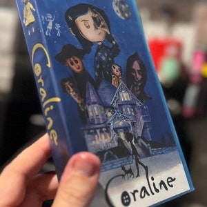 Może przedstawiać: Niebieskie pudełko z grafiką z filmu "Koralina i tajemnicze drzwi". Grafika na pudełku przedstawia postacie z filmu, w tym Koralinę, Drugą Matkę i dom. Tytuł "Koralina" jest napisany stylizowaną czcionką.