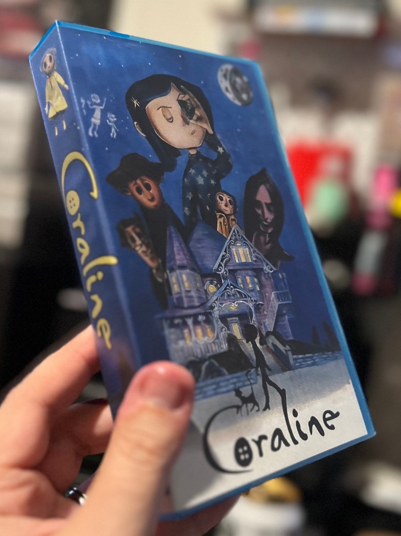 Może przedstawiać: Niebieskie pudełko z grafiką z filmu "Koralina i tajemnicze drzwi". Grafika na pudełku przedstawia postacie z filmu, w tym Koralinę, Drugą Matkę i dom. Tytuł "Koralina" jest napisany stylizowaną czcionką.