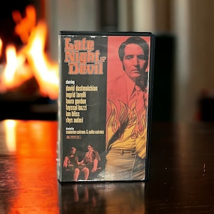 May include: A VHS tape cover for the movie "Late Night with the Devil". The cover features a man in a red and yellow shirt with flames behind him. The text "Late Night with the Devil" is in large, bold letters. The movie stars David Dastmalchian, Ingrid Torelli, Laura Gordon, Fayssal Bazzi, Ian Bliss, and Rhys Autery. It was directed by Cameron Cairnes and Colin Cairnes.