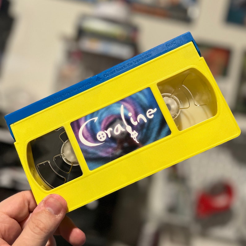 Może przedstawiać: Ż&oacute;łta kaseta VHS z niebieskim wierzchem. Na taśmie znajduje się tytuł filmu "Coraline" w białym skrypcie na wirującym, kolorowym tle. Taśma jest trzymana w dłoni.