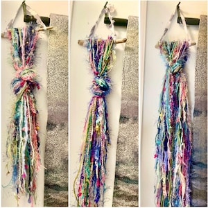 May include: A wall hanging composed of various multicolored yarn strands, ribbons, and textured fibers tied to a horizontal wooden branch. The hanging features a central knot and long, loose, dangling strands in shades of pink, blue, green, and yellow.