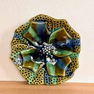 May include: A ceramic flower-shaped wall hanging with a glossy finish. The petals are shades of green and blue, with a textured, dotted border in a golden-brown hue. The center has a cluster of small white dots.