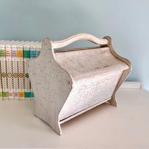 May include: A distressed white wooden magazine rack with a handle. The rack has a cracked paint finish and is sitting on a white surface. A row of books is visible in the background.