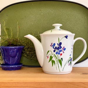 May include: A white ceramic teapot with a lid, adorned with blue and purple floral designs and green leaves. A small blue ceramic pot with a tulip design and a green tray are also present. The items are on a wooden surface.