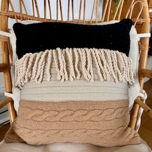 May include: A decorative throw pillow with a black, beige, and brown color scheme. The pillow has a fringe trim and a cable knit pattern.