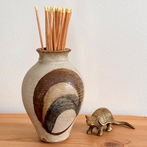 May include: A ceramic vase with a brown and tan swirl design holds a bundle of wooden matches. A small, brass armadillo figurine sits nearby. The vase and figurine are displayed on a wooden surface.