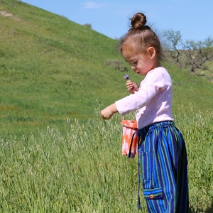 May include: A young child in a field of tall grass, wearing a light pink long-sleeved shirt and blue striped pants. The child is holding a purple flower and a small orange and white bucket. Green hills and a blue sky are in the background.