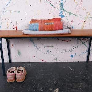 May include: A wooden bench with dark blue legs holds a stack of colourful textiles. The textiles include a red and orange patterned piece, a blue and white striped piece, and a light blue blanket. Two pairs of pink and tan shoes sit on the floor.
