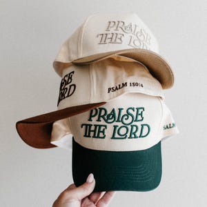 Op de afbeelding: Een stapel baseballpetten met de tekst "Praise the Lord" erop geborduurd. De petten zijn in verschillende kleuren, waaronder beige, bruin en groen. De tekst "Psalm 150:6" is ook zichtbaar op een van de petten.
