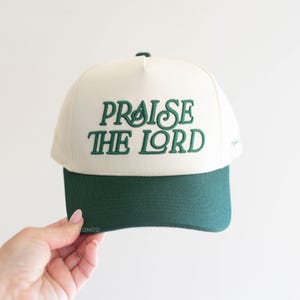 Op de afbeelding: Cr&egrave;mekleurige baseballcap met een donkergroen vizier. De tekst "PRAISE THE LORD" is in donkergroen op de voorkant geborduurd. De cap wordt tegen een witte achtergrond gehouden.