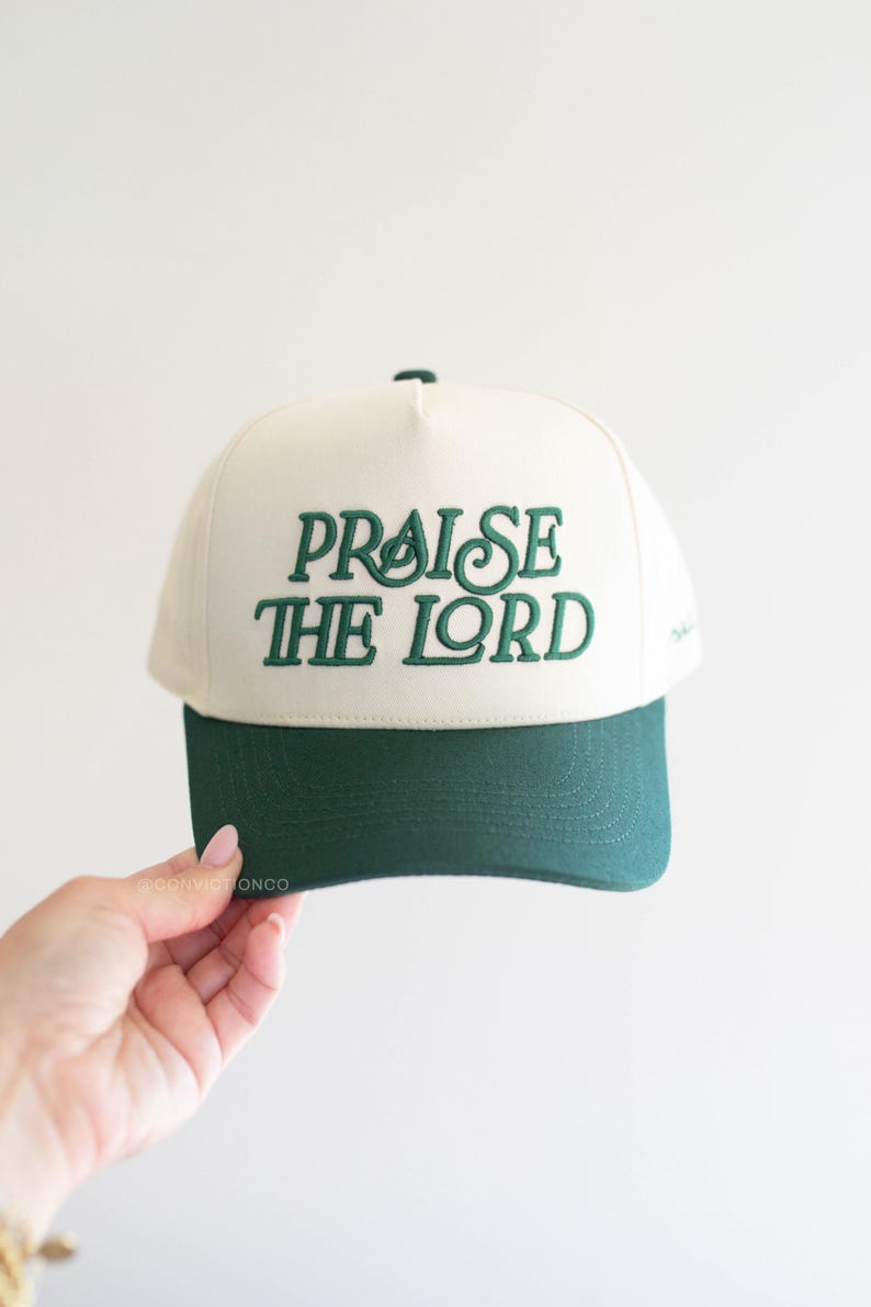 Op de afbeelding: Cr&egrave;mekleurige baseballcap met een donkergroen vizier. De tekst "PRAISE THE LORD" is in donkergroen op de voorkant geborduurd. De cap wordt tegen een witte achtergrond gehouden.