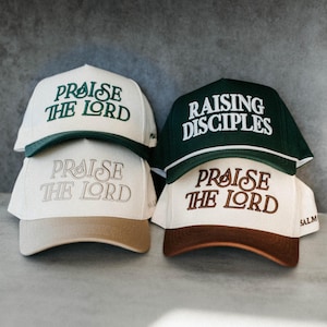 Op de afbeelding: Vier baseballpetten op elkaar gestapeld. Twee petten zijn wit met groene tekst die "Praise the Lord" luidt. E&eacute;n pet is donkergroen met witte tekst die "Raising Disciples" luidt. De andere twee petten zijn beige en bruin met de tekst "Praise the Lord".