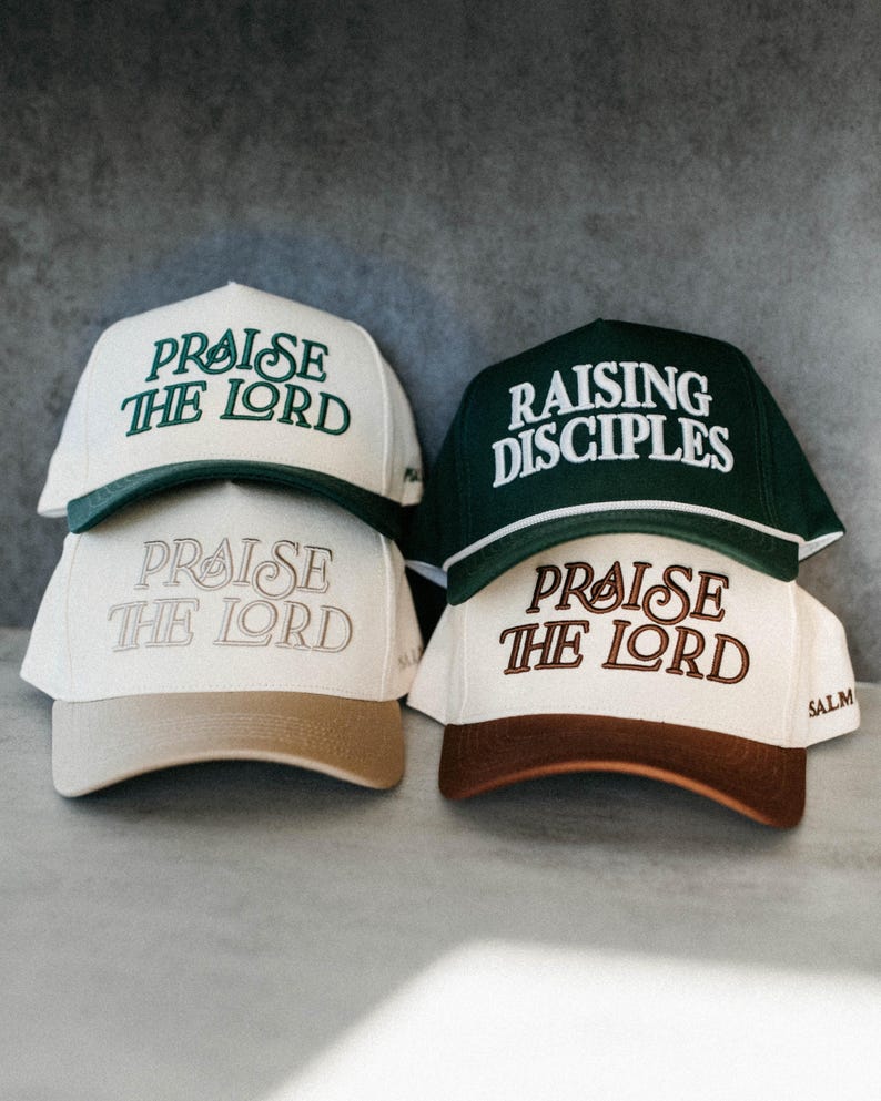 Op de afbeelding: Vier baseballpetten op elkaar gestapeld. Twee petten zijn wit met groene tekst die "Praise the Lord" luidt. E&eacute;n pet is donkergroen met witte tekst die "Raising Disciples" luidt. De andere twee petten zijn beige en bruin met de tekst "Praise the Lord".
