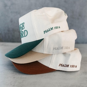 Op de afbeelding: Een stapel baseballpetten in cr&egrave;me, beige en bruin. De petten hebben donkergroene, beige en bruine klepjes. De tekst "PRAISE LORD" is op de bovenste pet geborduurd en "PSALM 150:6" is op elke pet geborduurd.