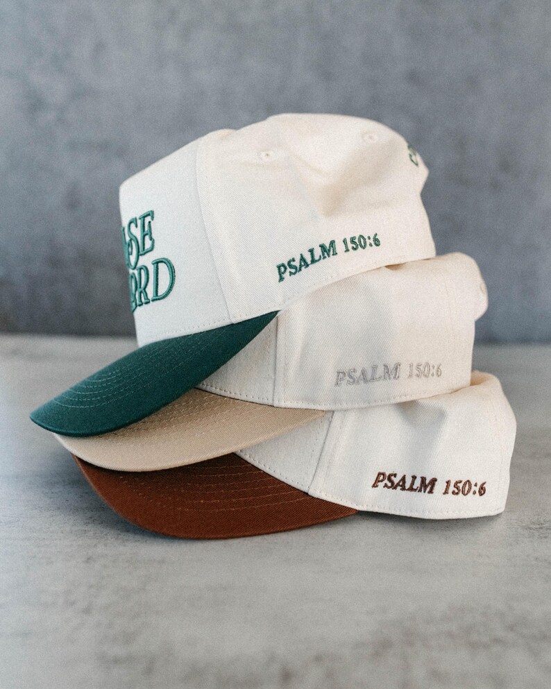 Op de afbeelding: Een stapel baseballpetten in cr&egrave;me, beige en bruin. De petten hebben donkergroene, beige en bruine klepjes. De tekst "PRAISE LORD" is op de bovenste pet geborduurd en "PSALM 150:6" is op elke pet geborduurd.