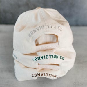 Op de afbeelding: Drie cr&egrave;mekleurige baseballpetten op elkaar gestapeld. Elke pet heeft de woorden "CONVICTION CO" of "CONVICTION" op de achterkant geborduurd in grijs, groen en bruin. De petten hebben verstelbare snapback-sluitingen.
