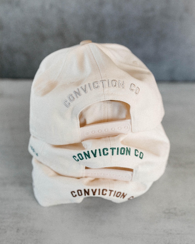 Op de afbeelding: Drie cr&egrave;mekleurige baseballpetten op elkaar gestapeld. Elke pet heeft de woorden "CONVICTION CO" of "CONVICTION" op de achterkant geborduurd in grijs, groen en bruin. De petten hebben verstelbare snapback-sluitingen.