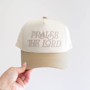 Op de afbeelding: Cr&egrave;mekleurige baseballcap met de geborduurde tekst "PRAISE THE LORD" in lichtbruin. De klep is effen lichtbruin van kleur. De pet wordt door een persoon vastgehouden.