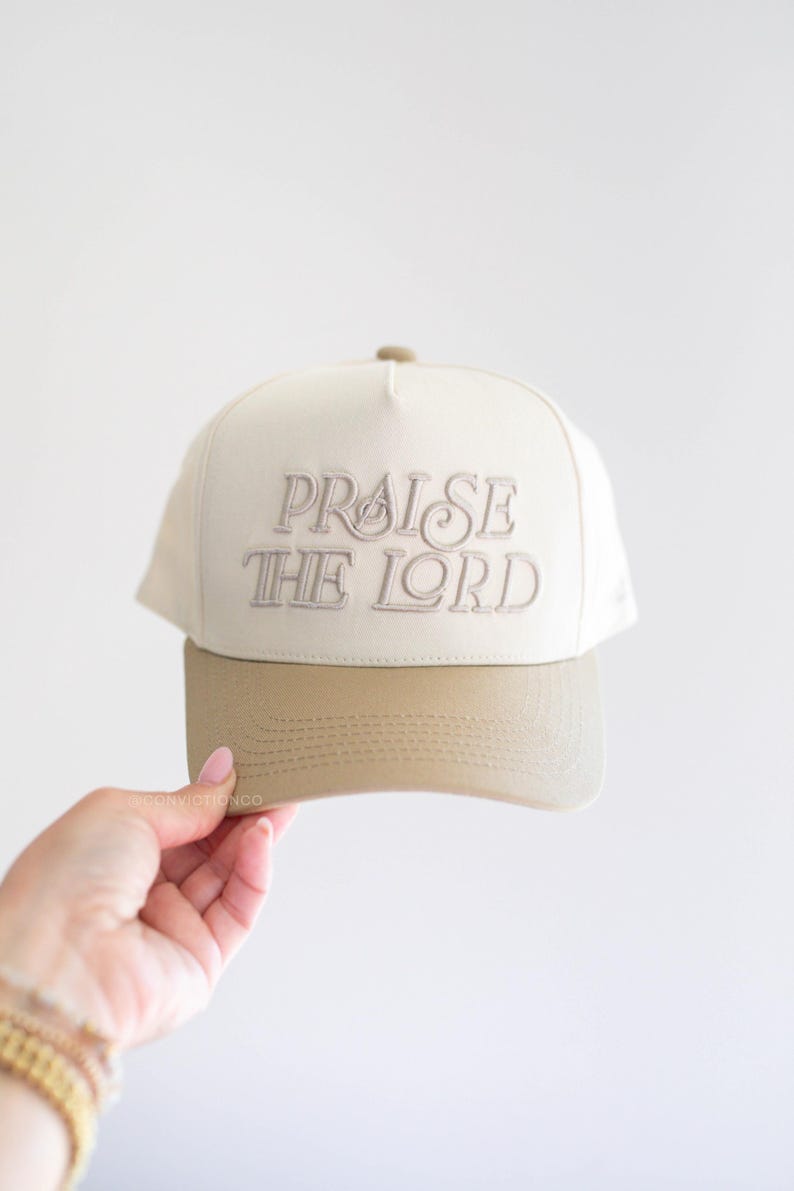 Op de afbeelding: Cr&egrave;mekleurige baseballcap met de geborduurde tekst "PRAISE THE LORD" in lichtbruin. De klep is effen lichtbruin van kleur. De pet wordt door een persoon vastgehouden.