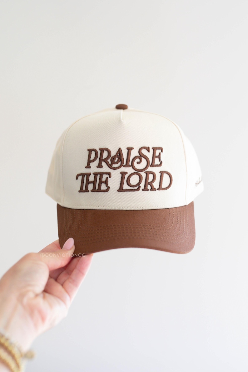 Op de afbeelding: Een baseballpet met een cr&egrave;mekleurige kroon en een bruine klep. De woorden "PRAISE THE LORD" zijn in bruin op de voorkant geborduurd. De pet heeft een bruine knop bovenop.