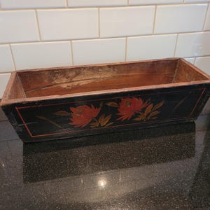 May include: Rectangular wooden planter box with a black exterior and red floral design. The box has a distressed appearance, with visible wear and tear on the wood. The floral design features red flowers and green leaves.