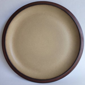 May include: A round, ceramic plate with a speckled tan center and a dark brown rim. The plate has a simple, minimalist design, suitable for serving meals. The plate is a neutral color, making it versatile for various table settings.