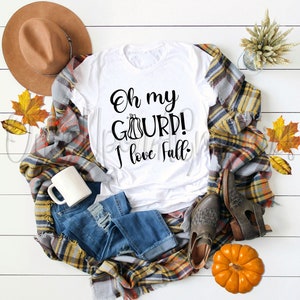 Könnte beinhalten: Weißes T-Shirt mit dem Text "Oh my gourd! I love fall." in schwarzer Schrift. Das Shirt liegt auf einer karierten Decke mit einem braunen Hut, einer Tasse, einem Paar braunen Stiefeln und einem Kürbis.