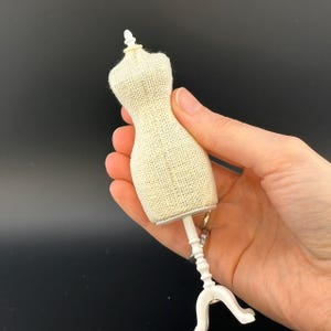 May include: A miniature dress form with a beige burlap-like fabric torso and a white stand. The dress form is designed to be a small-scale model, likely for sewing or fashion design purposes. The stand has a tripod base.