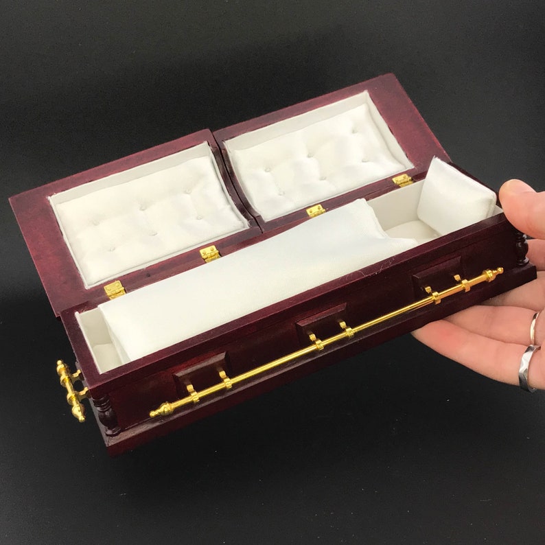 May include: A small, red wooden coffin with a gold metal handle and white satin lining. The coffin is open and the lining is visible.