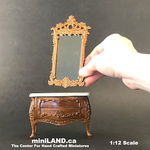 Puede incluir: Una cómoda de madera en miniatura con dos cajones y tallas ornamentadas. La cómoda está pintada de color marrón oscuro y tiene una tapa de mármol blanco. Un espejo dorado ornamentado en miniatura flota sobre la cómoda. El texto "miniLAND.ca The Center For Hand Crafted Miniatures 1:12 Scale" está en la parte inferior de la imagen.