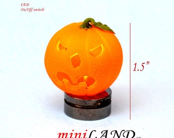 Può includere: Una zucca arancione in miniatura a forma di jack-o'-lantern con un viso intagliato e luci LED. La zucca è alta 3,8 cm e si trova su una base in metallo nero. Il testo "LED On/Off switch" è visibile nell'immagine.