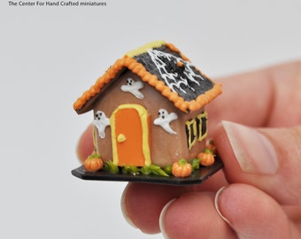 Może przedstawiać: Miniaturowy domek z piernika ozdobiony na Halloween. Domek jest brązowy z pomarańczowymi i żółtymi akcentami. Dach jest czarny z białym wzorem pajęczyny. Na froncie domku znajdują się trzy białe duchy. Domek stoi na czarnej podstawie z zieloną trawą i dwiema pomarańczowymi dyniami.