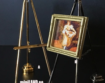 May include: Two miniature easels, one gold and one black, with a small framed painting on the black easel. The painting depicts a woman in a white dress holding a basket of flowers. The easels are 1:12 scale.