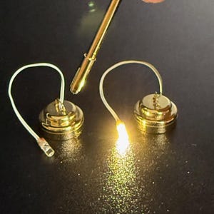 Puede incluir: Dos pequeñas luces LED doradas con un brillo cálido, conectadas por finos cables blancos. Las luces son redondas con una pequeña base cilíndrica. También hay una herramienta de metal dorado en la imagen, posiblemente para la instalación o el uso.