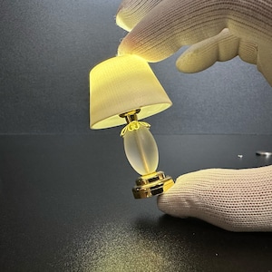 May include: A small lamp featuring a cream lampshade and a gold base. The lamp has a frosted glass centre and is lit, emitting a soft light. The lamp is held by a gloved hand against a dark backdrop.