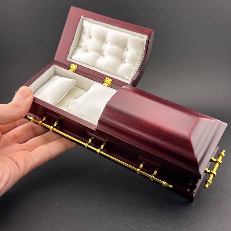 May include: A small, wooden coffin-shaped box with a gold trim and a velvet lining. The lid is open, revealing the interior.