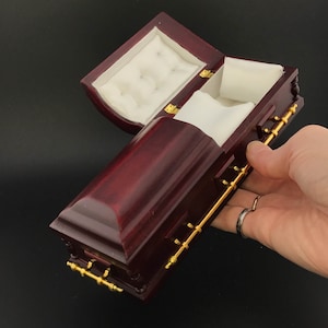 May include: A small, dark red wooden coffin-shaped box with gold accents and a white velvet interior. The lid is open, revealing the empty interior.