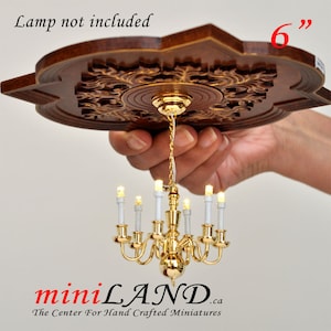 May include: A gold-toned miniature chandelier with white candle-shaped bulbs hangs from a wooden ceiling medallion. The medallion is 15 cm in diameter and has an ornate design. The chandelier is approximately 15 cm tall. "Lamp not included" is written above the medallion.