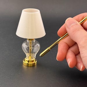 Pode incluir: Uma lâmpada de mesa de ouro em miniatura com um abajur de tecido branco e uma base de vidro transparente. A lâmpada está sendo segurada por uma mão com uma haste de metal dourado.